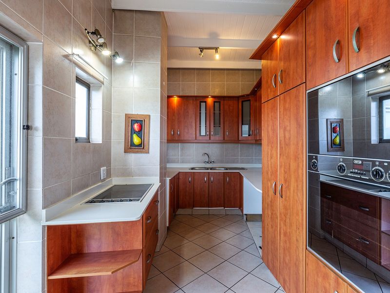 A well-maintained kitchen featuring warm-toned wooden cabinetry, tiled flooring, integrated oven, electric hob, and ample counter space with modern lighting.