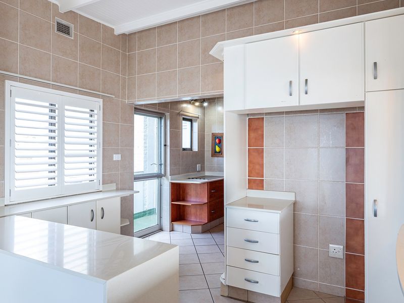 A clean, well-lit kitchen featuring neutral tiled walls, ample built-in cabinetry, stone countertops, and plantation shutters, with an adjacent scullery area.