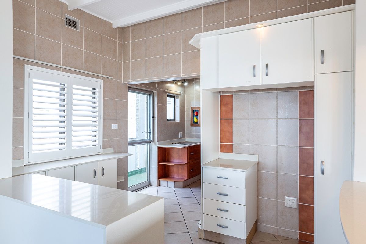 A clean, well-lit kitchen featuring neutral tiled walls, ample built-in cabinetry, stone countertops, and plantation shutters, with an adjacent scullery area.
