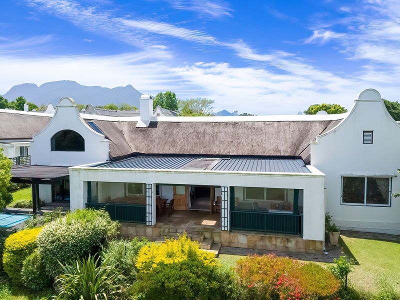 Rear view of a charming Cape Dutch style home featuring a thatched roof, a spacious covered patio, and a private swimming pool set in a garden.