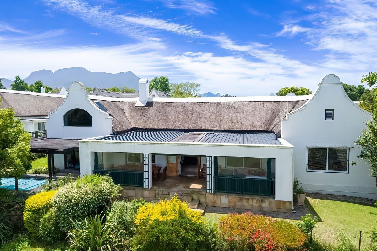 Rear view of a charming Cape Dutch style home featuring a thatched roof, a spacious covered patio, and a private swimming pool set in a garden.