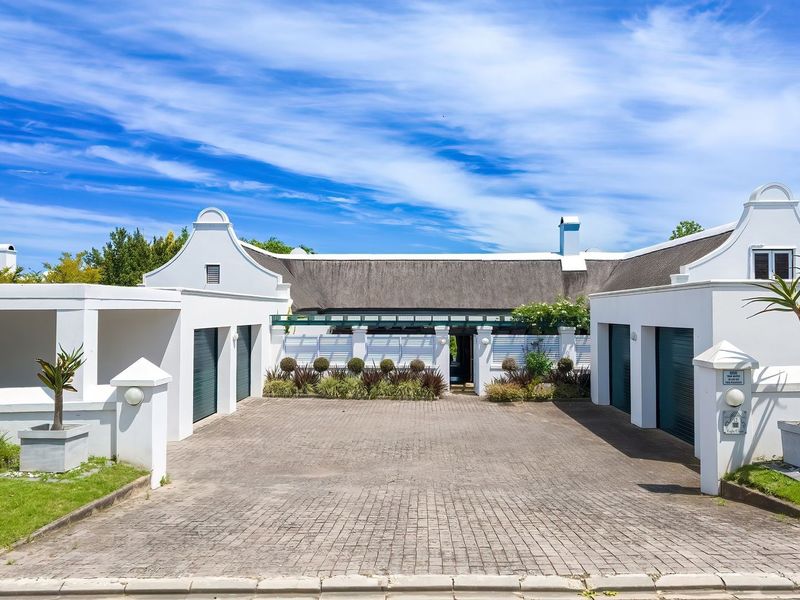 A well-maintained front elevation featuring a classic gabled facade, a thatched roof, and a spacious paved driveway leading to a double garage.