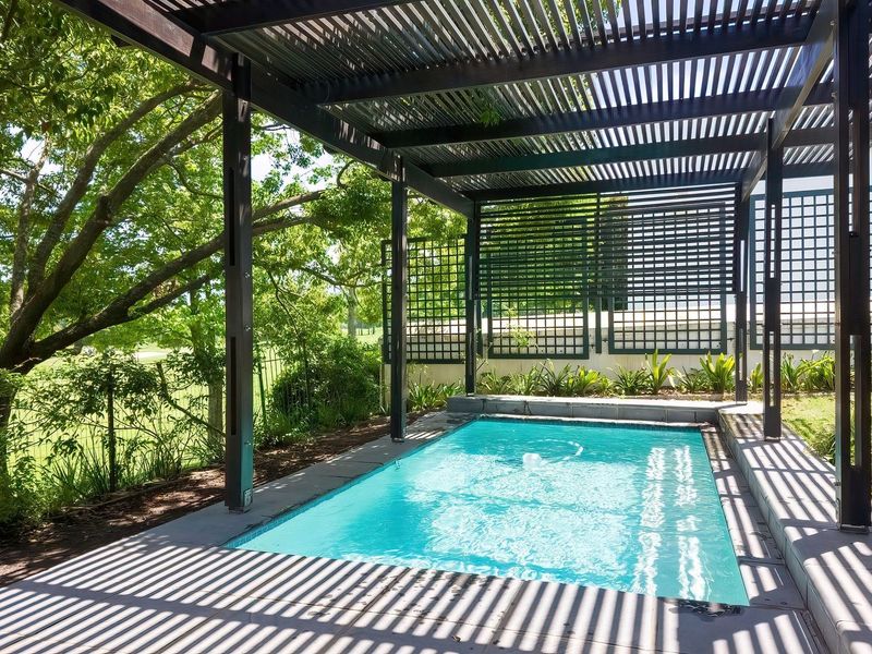 A well-maintained swimming pool area featuring a modern pergola structure, stone paving, and decorative trellis fencing, set within a lush garden environment.