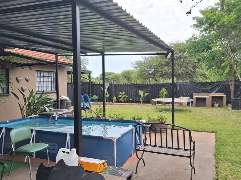 Covered patio area featuring a concrete slab, steel roofing structure, and a built-in outdoor braai, overlooking a garden with perimeter fencing.