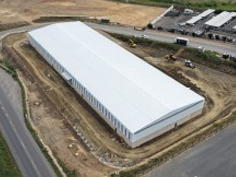 Aerial view of a large industrial warehouse building situated on a corner plot with surrounding access roads and undeveloped grounds.