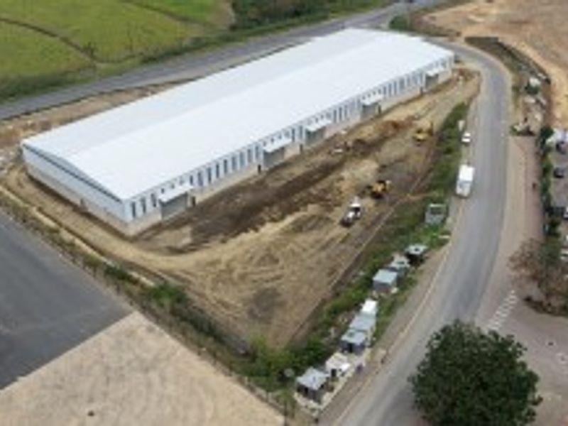 Aerial view of a large industrial warehouse facility under development, featuring a metal roof, paved access roads, and surrounding undeveloped land.