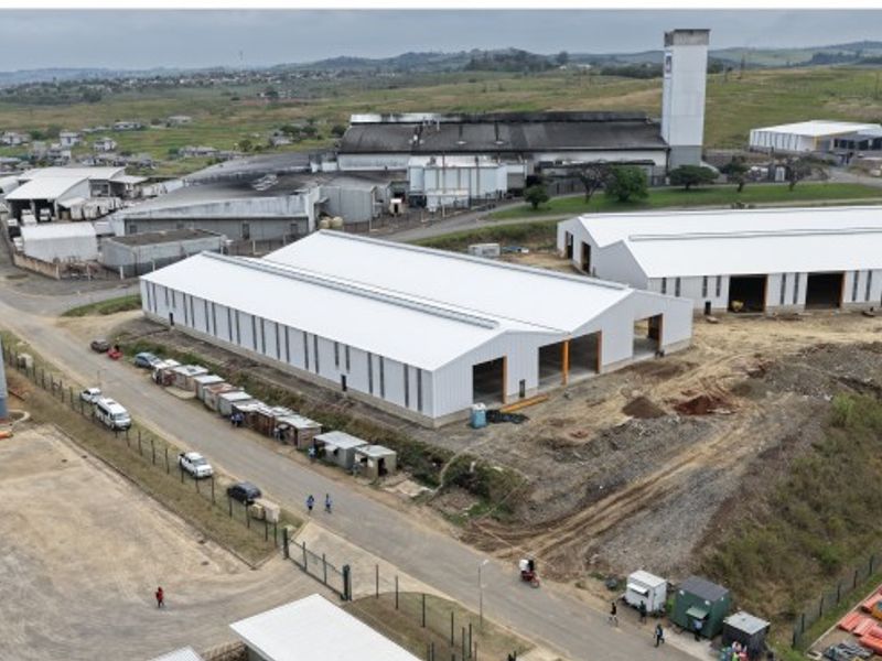 Aerial view of an industrial facility featuring large warehouse structures with metal roofing, perimeter fencing, and adjacent water storage tanks.