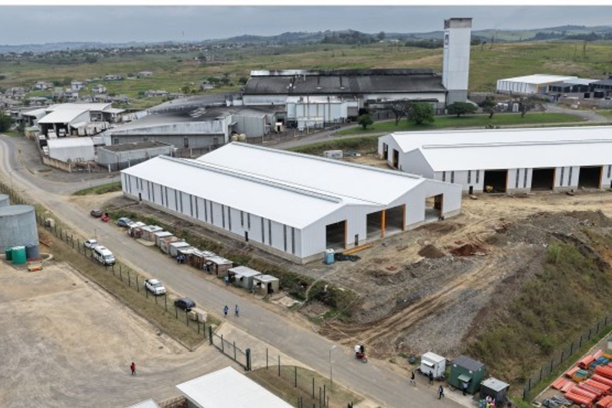Aerial view of an industrial facility featuring large warehouse structures with metal roofing, perimeter fencing, and adjacent water storage tanks.