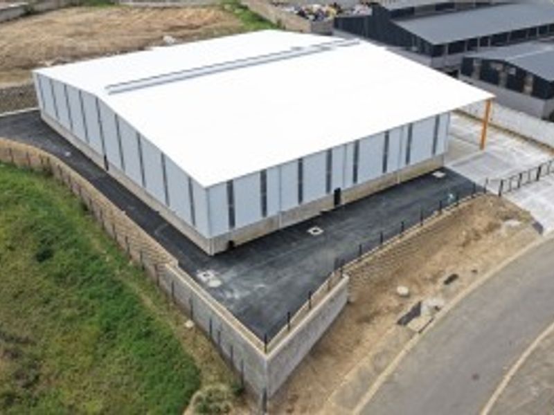 Aerial view of a modern industrial warehouse facility featuring a large metal-clad structure, paved driveway, and secure perimeter fencing.