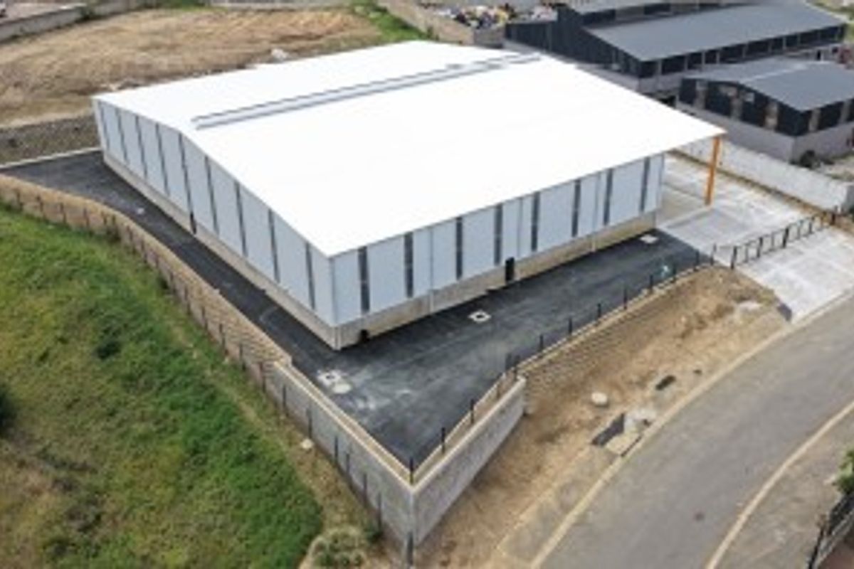 Aerial view of a modern industrial warehouse facility featuring a large metal-clad structure, paved driveway, and secure perimeter fencing.
