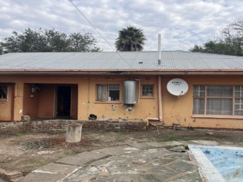 Rear view of the property featuring a corrugated metal roof, swimming pool, and stone patio area requiring maintenance and exterior refurbishment.
