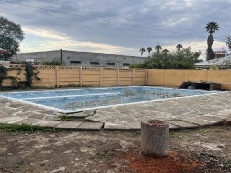 Outdoor pool area featuring a large swimming pool with surrounding stone paving and perimeter boundary walls, currently requiring significant maintenance and restoration.