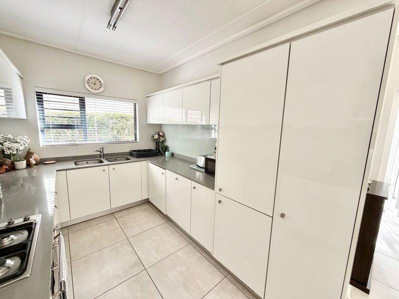 A modern, well-maintained kitchen featuring white high-gloss cabinetry, stone countertops, a double sink, and a built-in hob with tiled flooring.