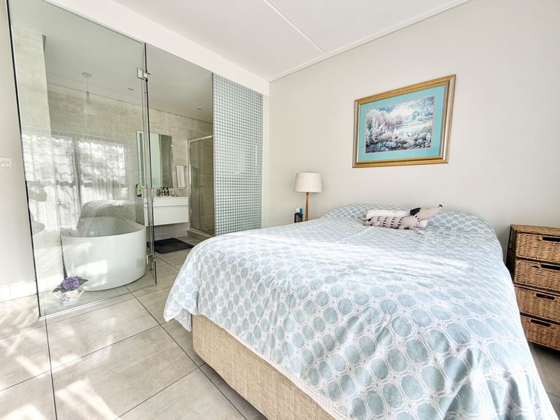 A modern bedroom featuring an open-plan en-suite bathroom with a glass partition, freestanding bathtub, walk-in shower, and contemporary tiled flooring.