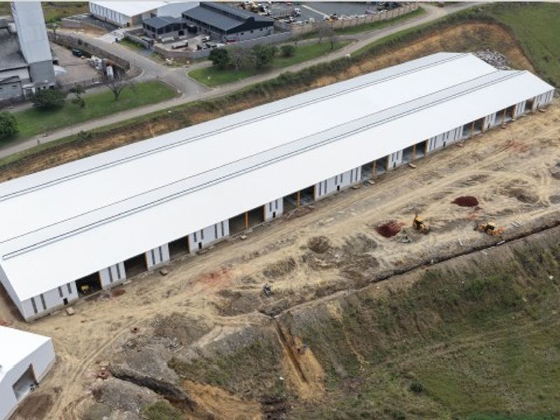 Aerial view of a large industrial warehouse facility under construction, featuring a long metal roof structure and surrounding undeveloped land.