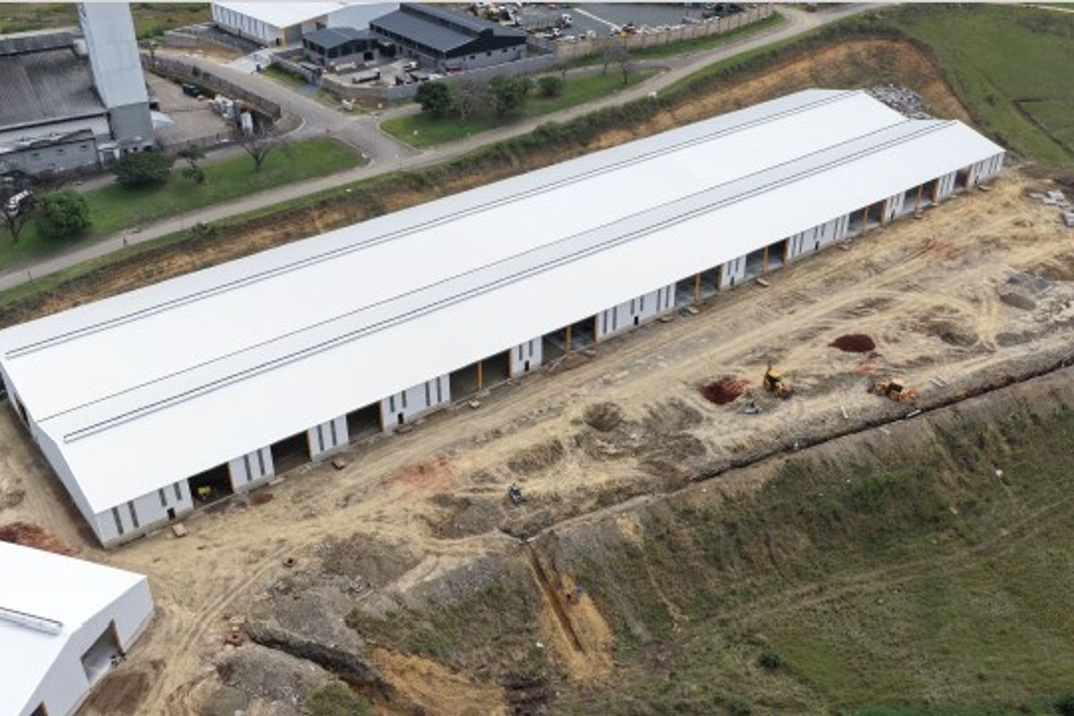 Aerial view of a large industrial warehouse facility under construction, featuring a long metal roof structure and surrounding undeveloped land.
