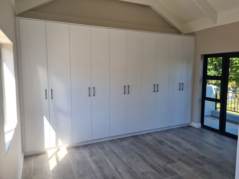 A bright, modern bedroom featuring extensive floor-to-ceiling built-in cupboards, vaulted ceilings, and durable timber-look tile flooring with access to an outdoor balcony.