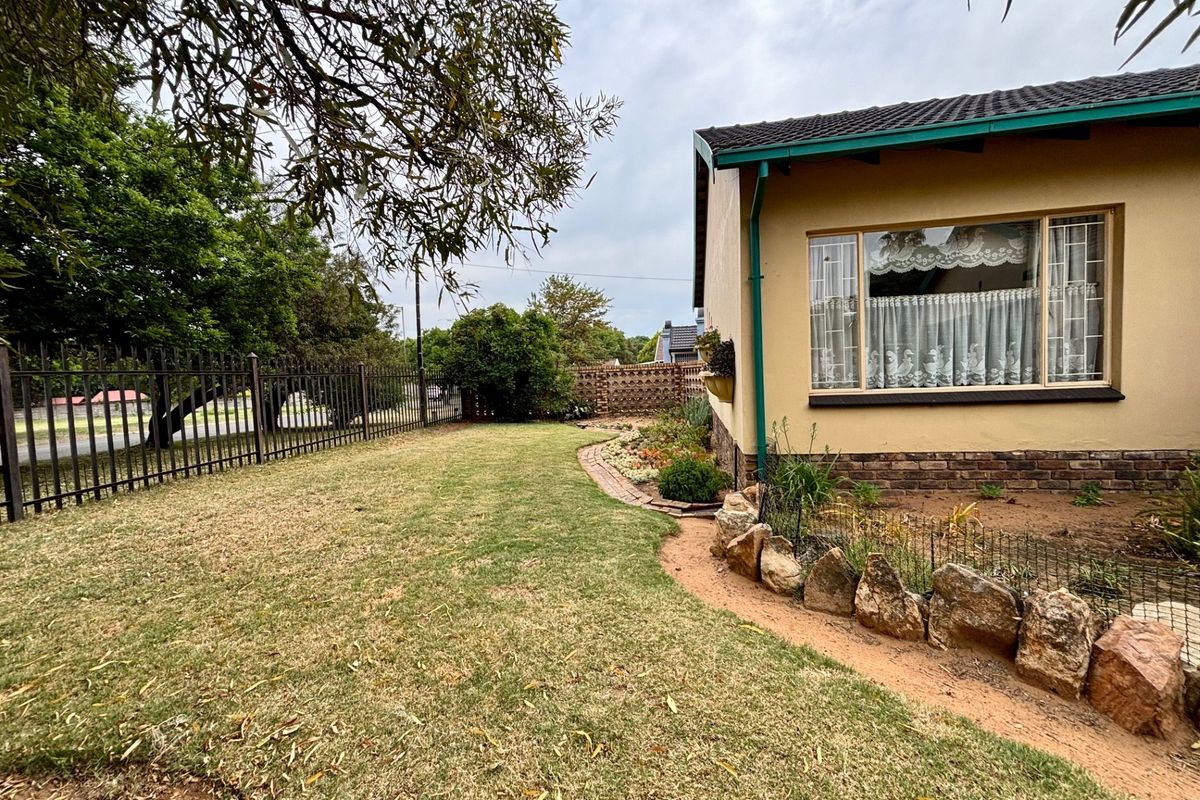 A view of the rear garden featuring a lawn, stone-edged garden beds, a brick path, and metal perimeter fencing alongside the house exterior.