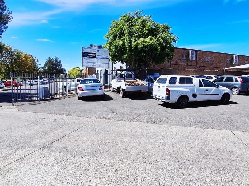 Exterior view of an industrial park entrance featuring an asphalt parking area, security fencing, and commercial signage on the building facade.