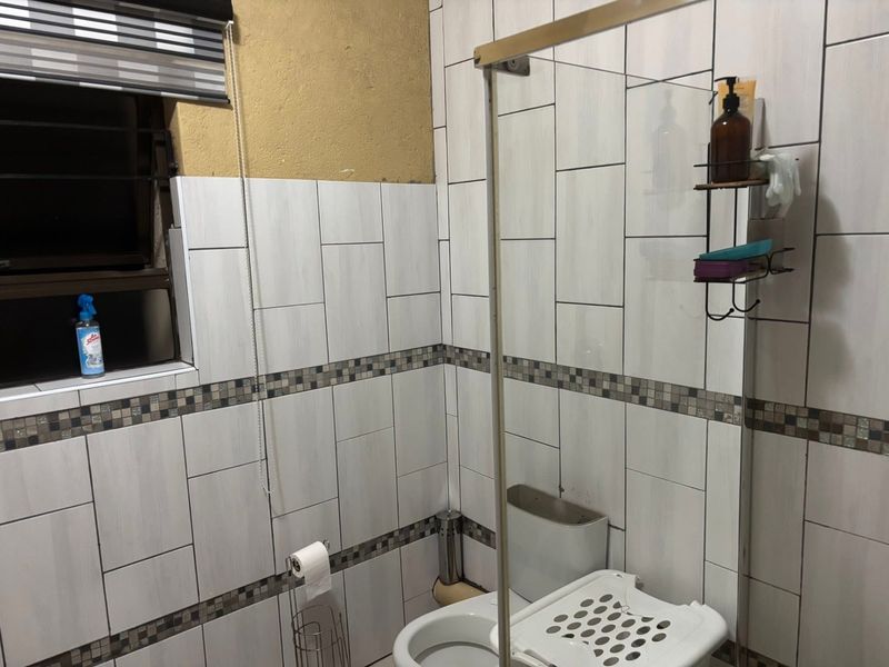 A bathroom featuring tiled walls with a mosaic border, a glass shower screen, and a toilet. The upper wall area requires finishing.