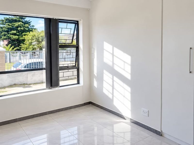 A bright, empty bedroom featuring modern tiled flooring, built-in cupboards, and a large window with security bars, offering a clean and neutral living space.