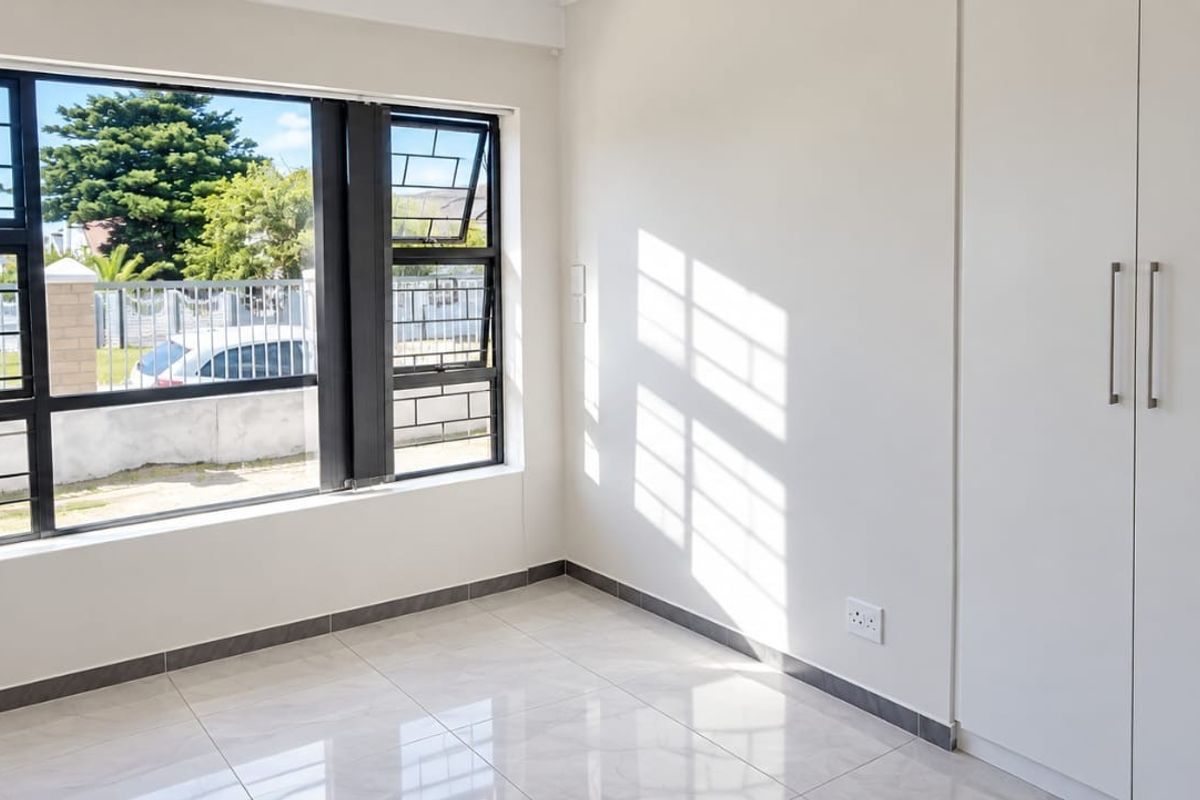 A bright, empty bedroom featuring modern tiled flooring, built-in cupboards, and a large window with security bars, offering a clean and neutral living space.