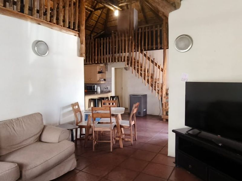 Open plan living and dining area featuring a high thatched ceiling, wooden staircase leading to a mezzanine level, and tiled flooring throughout.