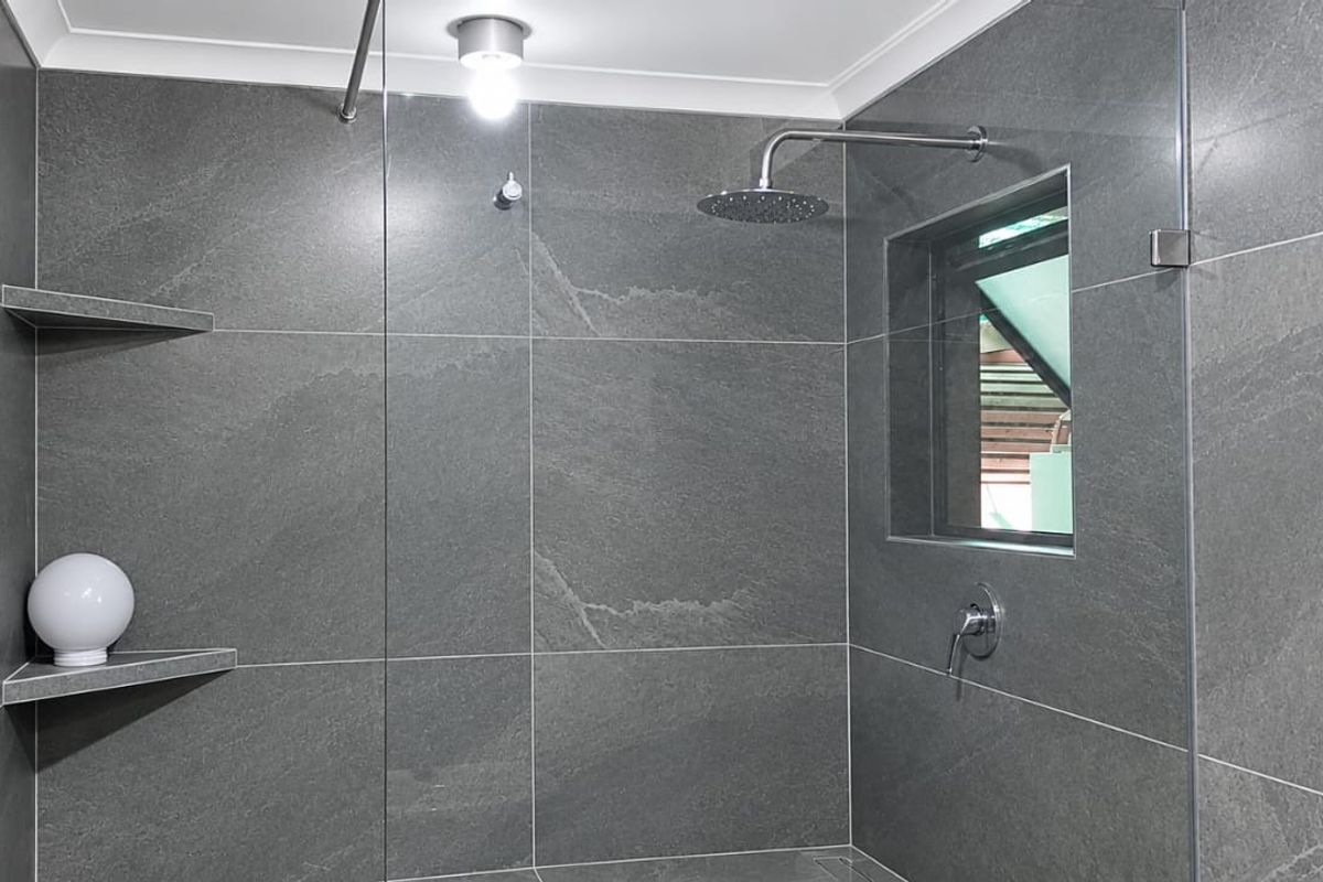 Modern walk-in shower featuring large-format grey stone-look tiles, a rain shower head, glass screen, and convenient built-in corner shelving.