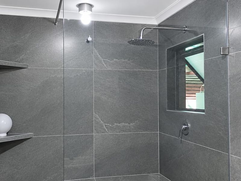 Modern walk-in shower featuring large-format grey stone-look tiles, a rain shower head, glass screen, and convenient built-in corner shelving.