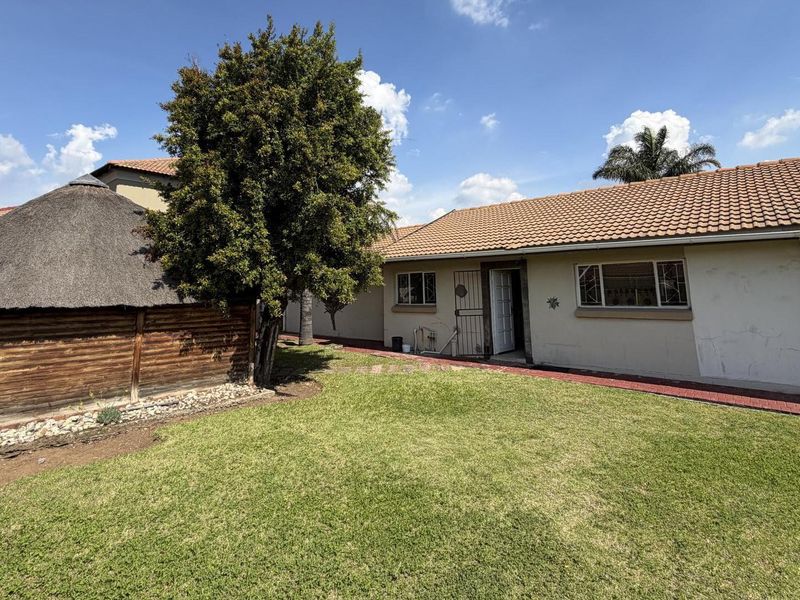 Rear view of the property featuring a lawn, paved walkway, and a thatched lapa structure adjacent to the main house.