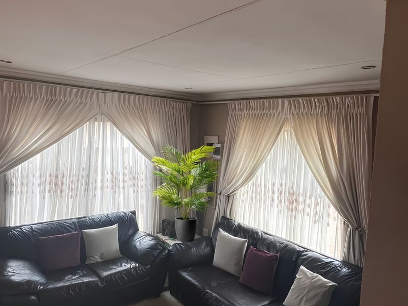 A comfortable living room featuring recessed downlights, decorative cornicing, and large windows dressed with elegant floor-to-ceiling curtains.
