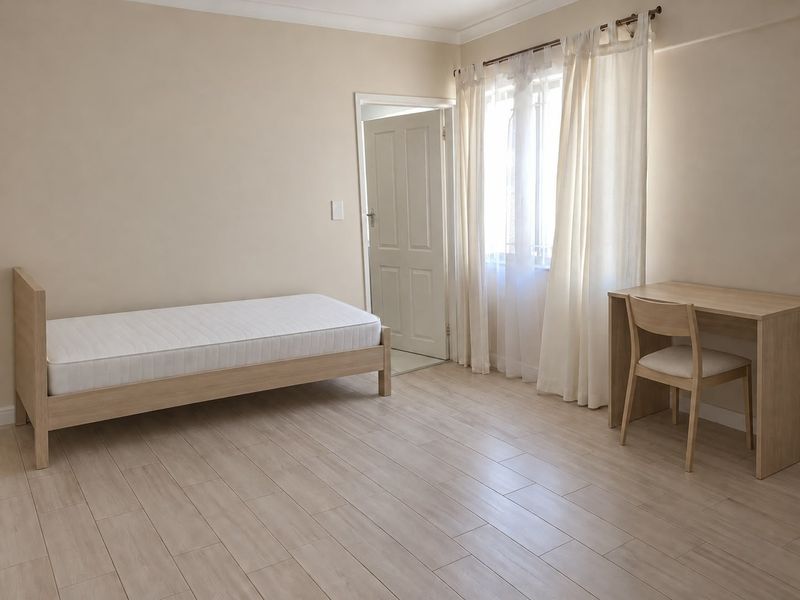 A bright and neutral bedroom featuring light-toned laminate flooring, a window with security bars, and a clean, minimalist aesthetic.