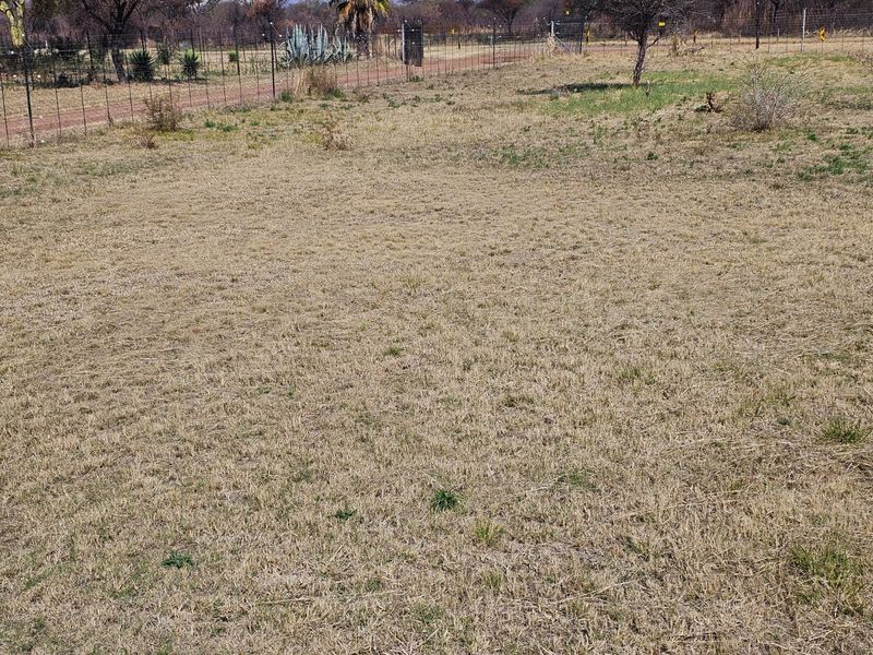 A view of a vacant residential plot featuring dry grass, perimeter wire mesh fencing, and a gated entrance.