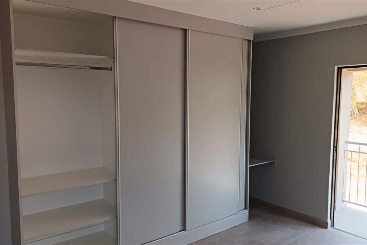 A modern bedroom featuring built-in sliding door wardrobes with integrated shelving and a small desk area, leading to a private balcony.