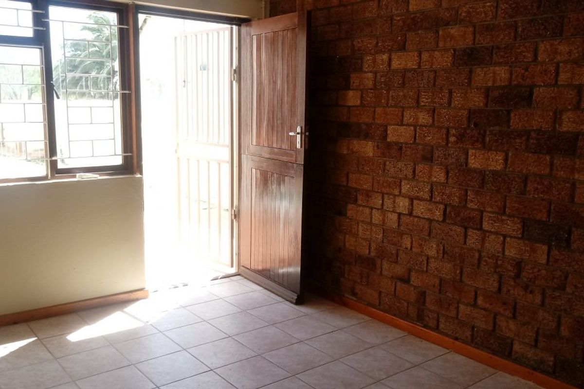 An empty living area featuring an exposed brick feature wall, tiled flooring, and direct access to the exterior through a timber door with security gate.