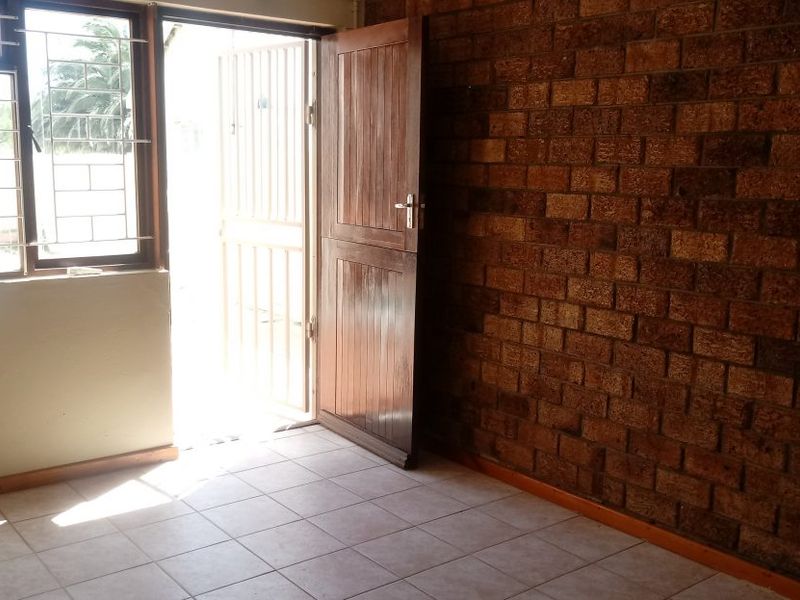 An empty living area featuring an exposed brick feature wall, tiled flooring, and direct access to the exterior through a timber door with security gate.