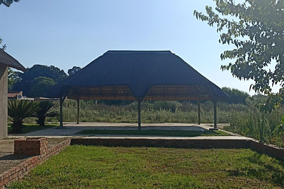 A spacious outdoor thatched-roof entertainment area set within a large garden, featuring a concrete base and open-sided structure.