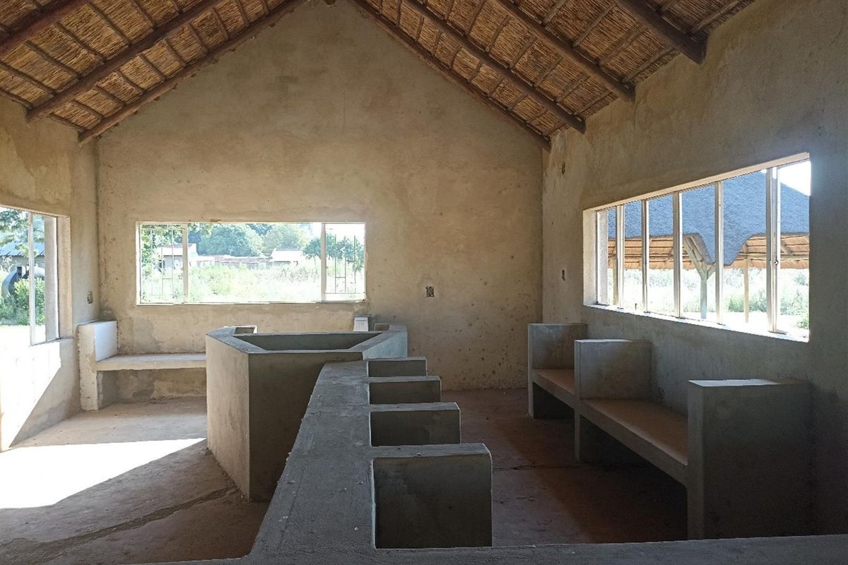 An open-plan interior space featuring a thatched roof, exposed concrete built-in structures, and large windows, currently in an unfinished shell condition.