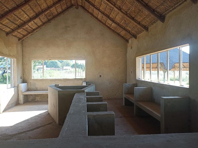 An open-plan interior space featuring a thatched roof, exposed concrete built-in structures, and large windows, currently in an unfinished shell condition.
