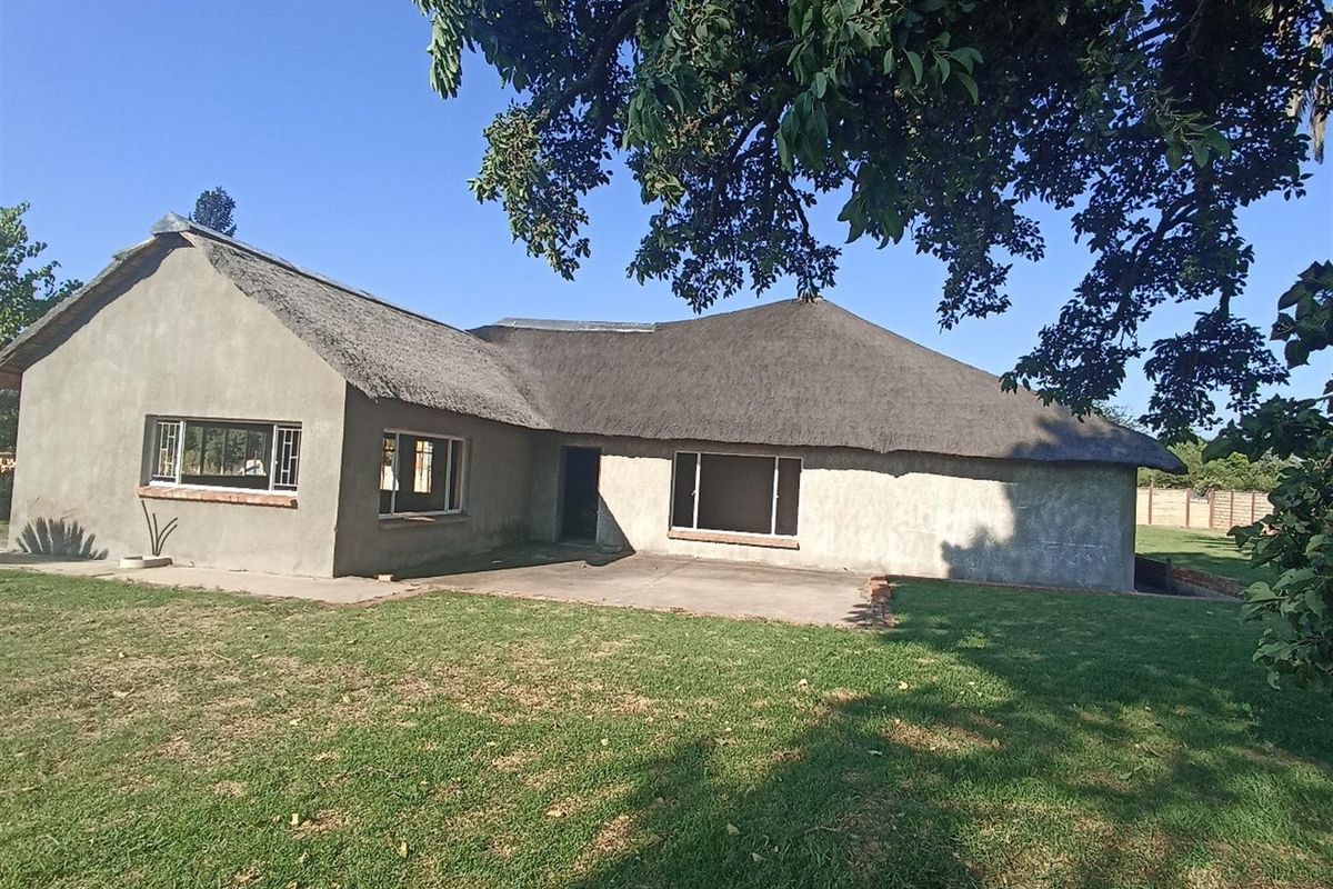Rear view of a farmhouse featuring a traditional thatched roof, concrete patio area, and surrounding lawn under mature trees.