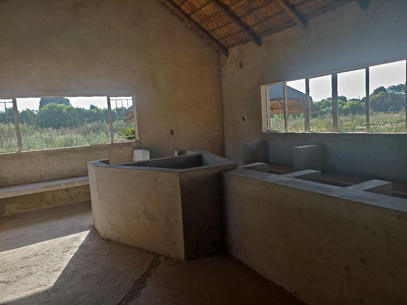 An unfinished interior space featuring exposed concrete structures, a thatched roof, and large window openings overlooking the surrounding farm landscape.