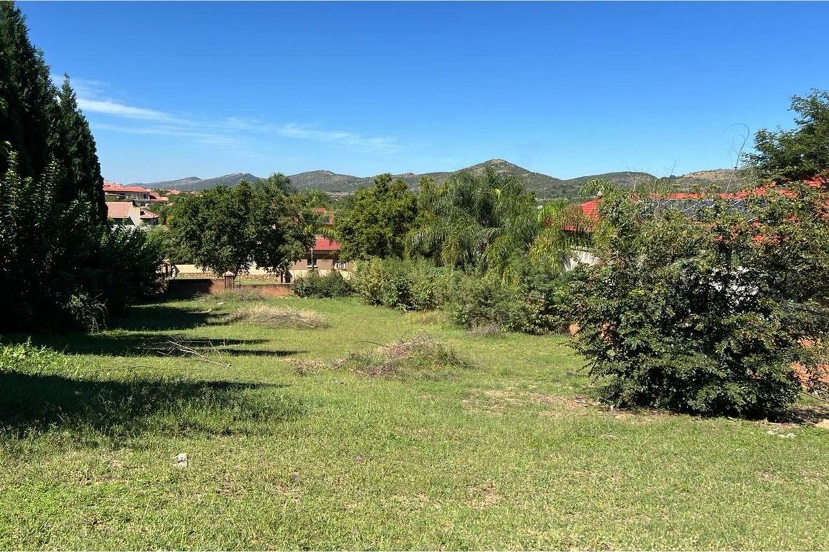 A view of a vacant residential plot featuring established trees and grass, with surrounding neighborhood homes and mountain views in the background.