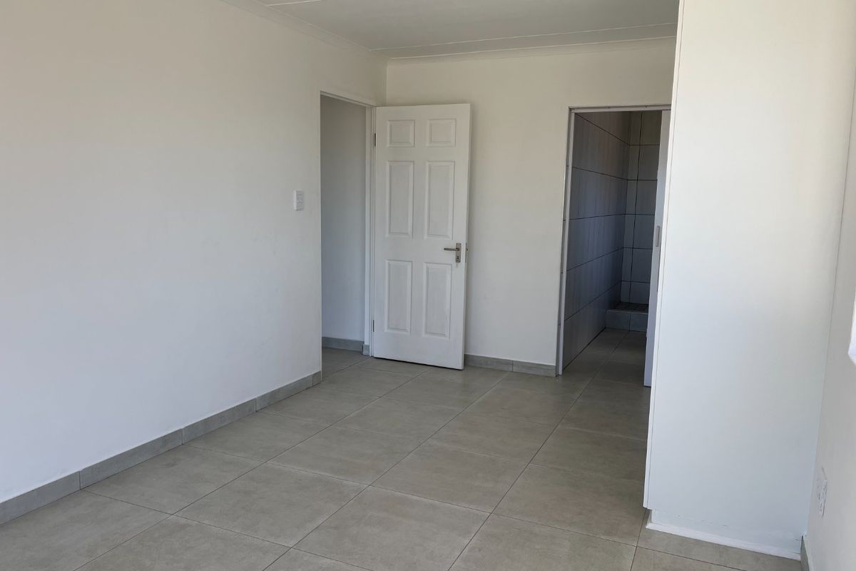 A bright, empty bedroom featuring neutral tiled flooring, clean white walls, and access to an adjoining bathroom area.
