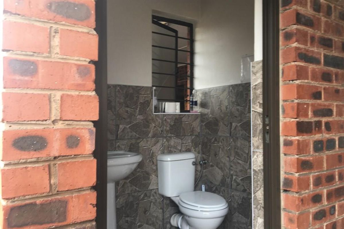 A compact bathroom featuring stone-look wall tiles, a pedestal basin, and a standard toilet, viewed through a brick-framed doorway.