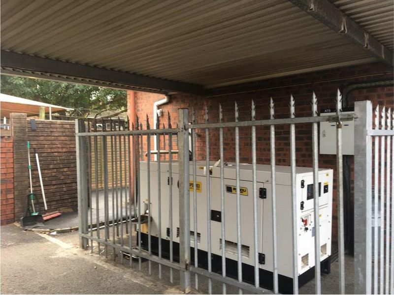 A secure outdoor utility area featuring a commercial-grade generator housed under a metal carport structure with perimeter security fencing.