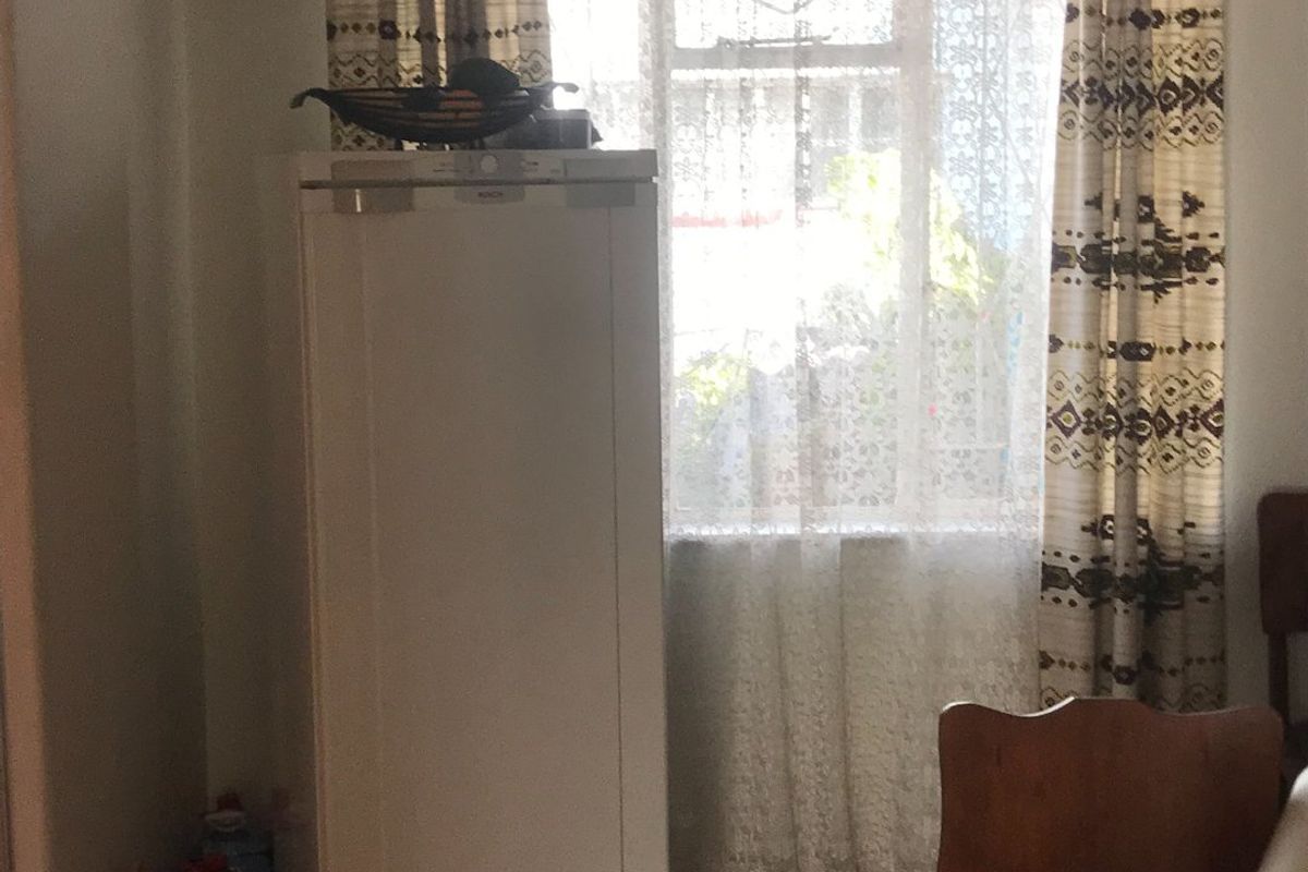 A view of a room featuring a window with security bars, lace curtains, and a ventilation grille, showing signs of age and wear.