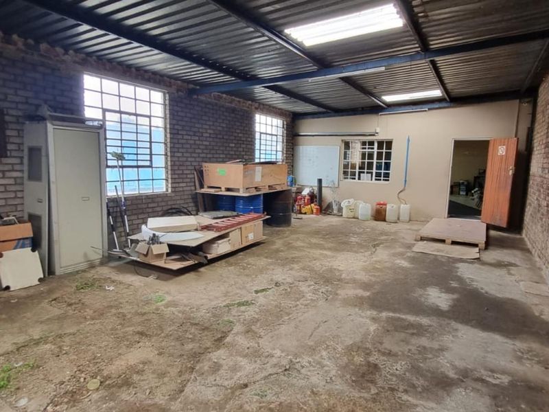 Industrial warehouse space featuring exposed brick walls, concrete flooring, and high-clearance corrugated metal roofing with integrated fluorescent lighting.