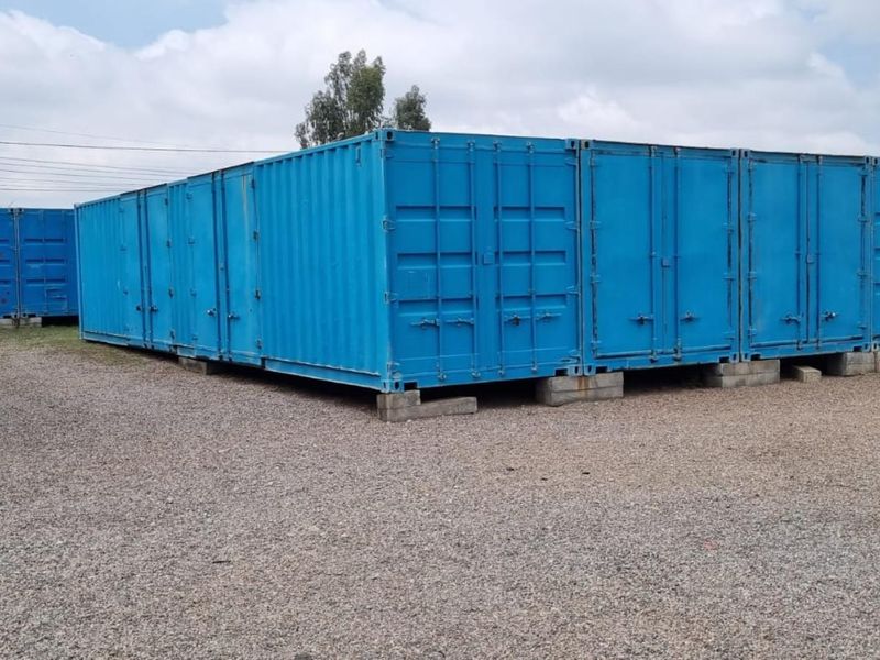 A series of industrial shipping containers used for storage, situated on a gravel yard surface.