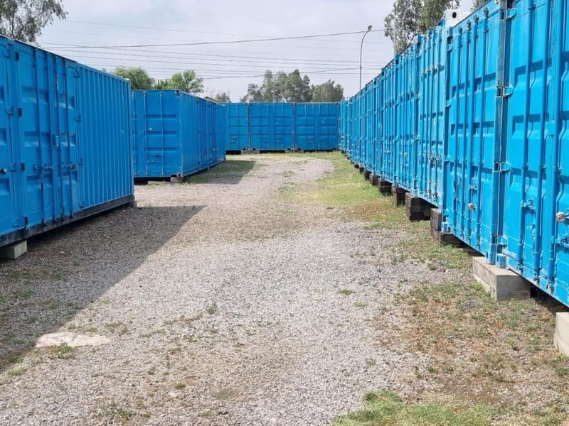 Industrial storage facility featuring multiple blue shipping containers arranged in rows on a gravel-surfaced yard.