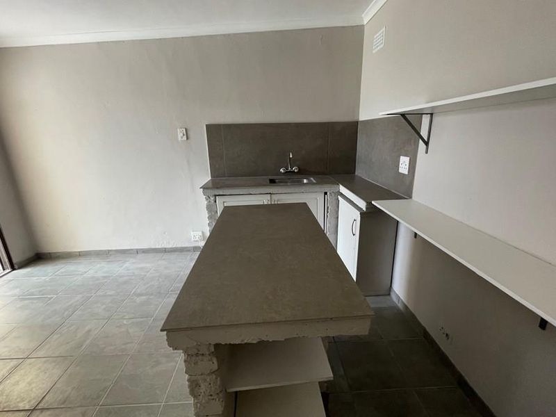 A basic kitchen area featuring a central island, tiled flooring, sink, and wall-mounted shelving, currently in need of cosmetic updates and finishing touches.